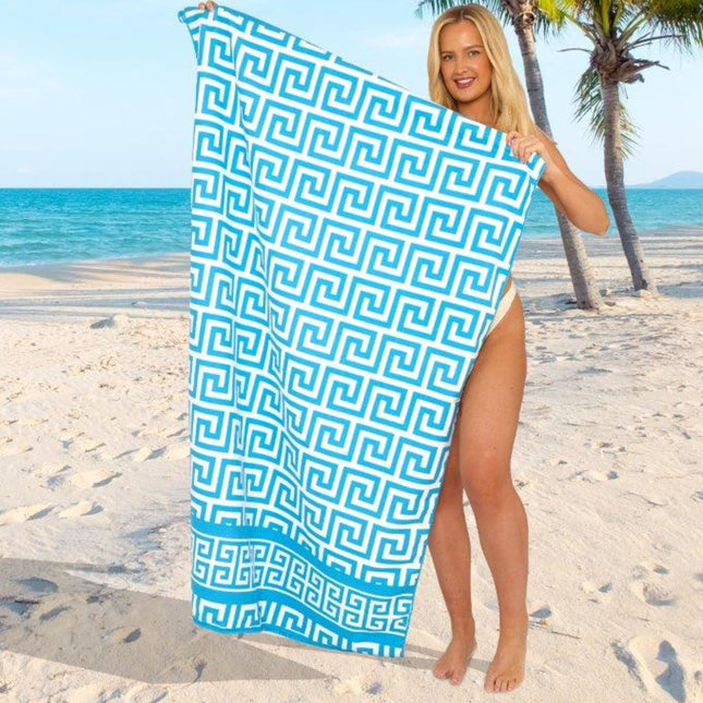 The image features a person standing on a sandy beach, holding a towel with a sophisticated Greek key pattern in blue and white. The interlocking geometric design gives the towel a classic and stylish look, perfect for a refined beach aesthetic. The backdrop includes the ocean and palm trees, reinforcing the tropical, relaxing vibe of the scene. This towel’s elegant motif makes it a standout choice for a seaside getaway or poolside lounging.