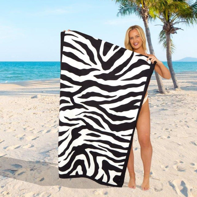 The image showcases a person standing on a sandy beach, holding a towel with bold zebra stripes in black and white. The striking contrast of the pattern makes this towel a standout piece, perfect for a stylish, adventurous beach day. The backdrop includes clear blue water, a sunny sky, and palm trees, enhancing the tropical, relaxed atmosphere. This towel’s wild yet elegant design is ideal for adding a touch of untamed charm to any seaside getaway!